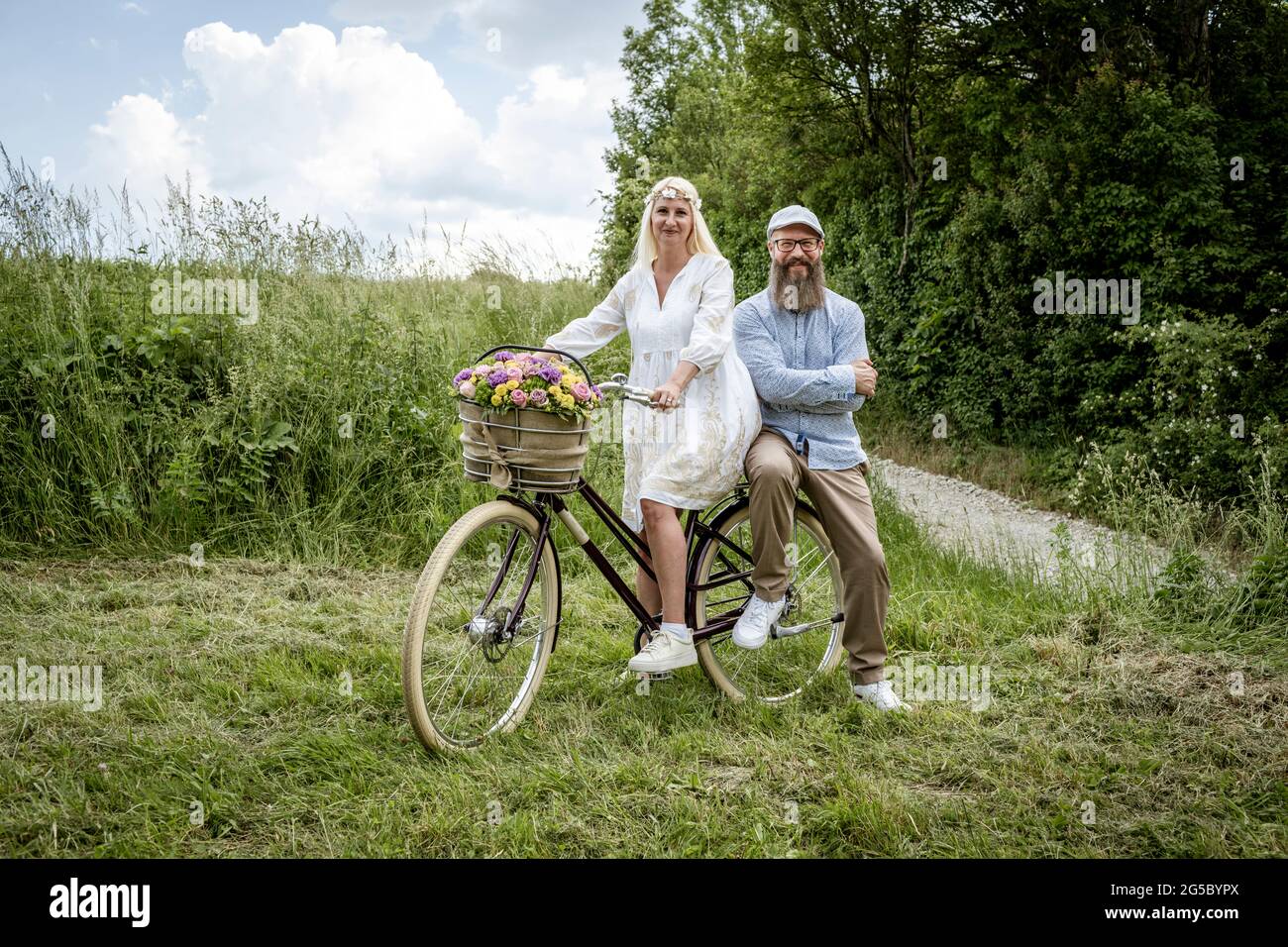 blonde woman with white dress and her boyfriend or husband posing with bicycle with beautifully decorated flower basket in nature and are happy and in Stock Photo