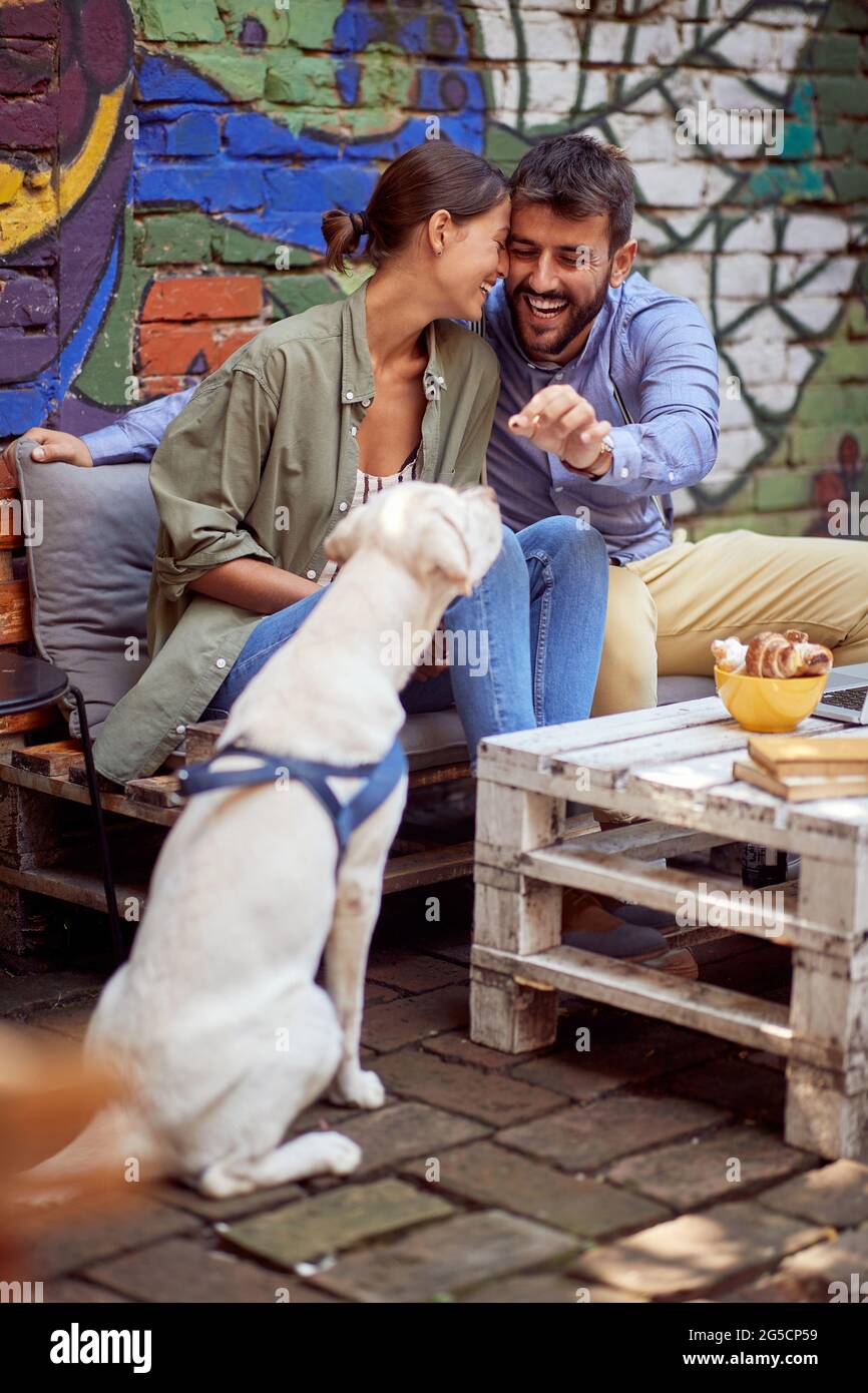 young couple having fun with their dog in outdoor cafe, cuddling each other and feeding pet, labrador retriever. Stock Photo