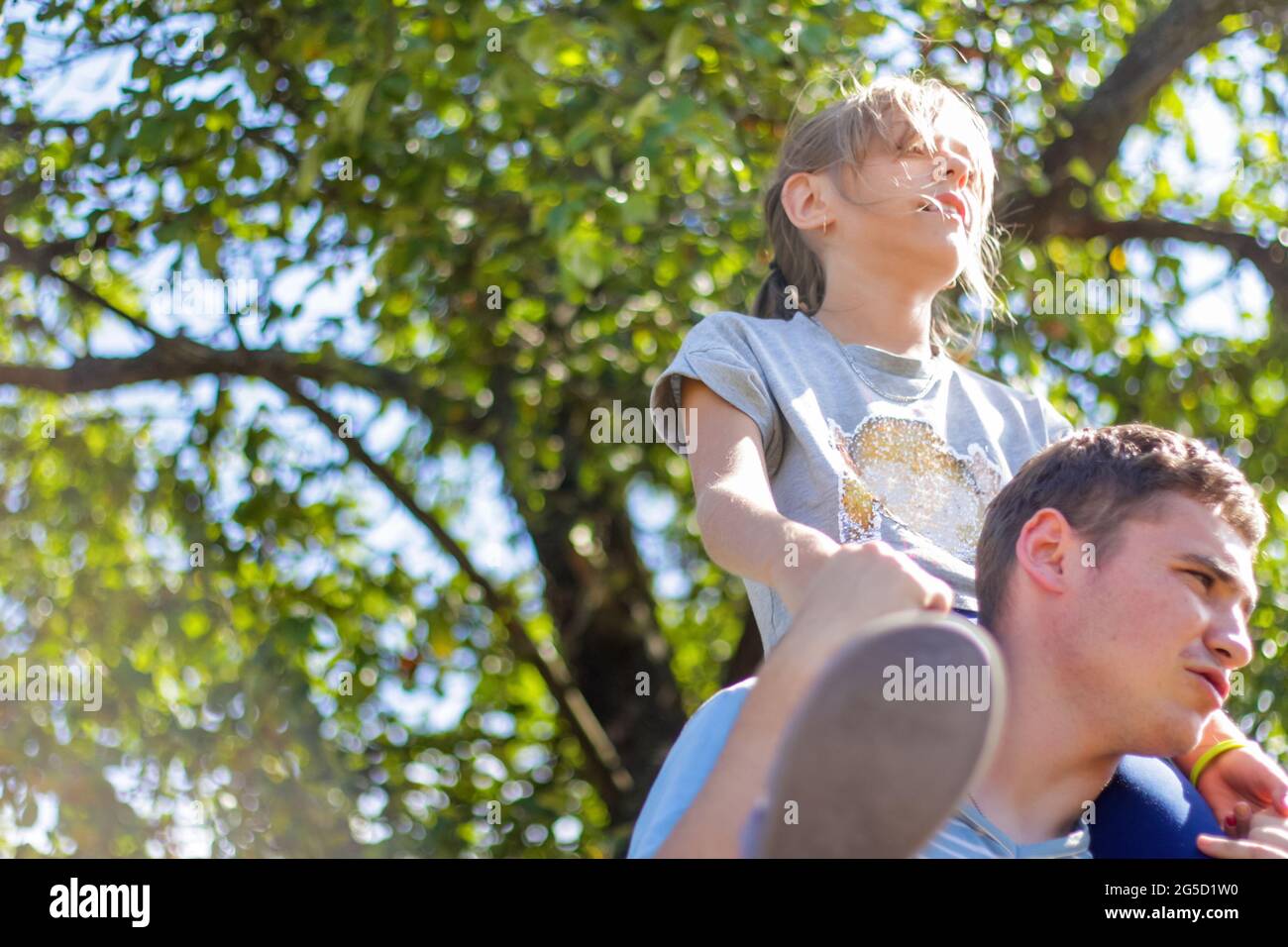Defocused brother riding sister on back. Portrait of happy girl on man shoulders, piggyback. Girl fly. Family playing outside outdoor. Green tree Stock Photo