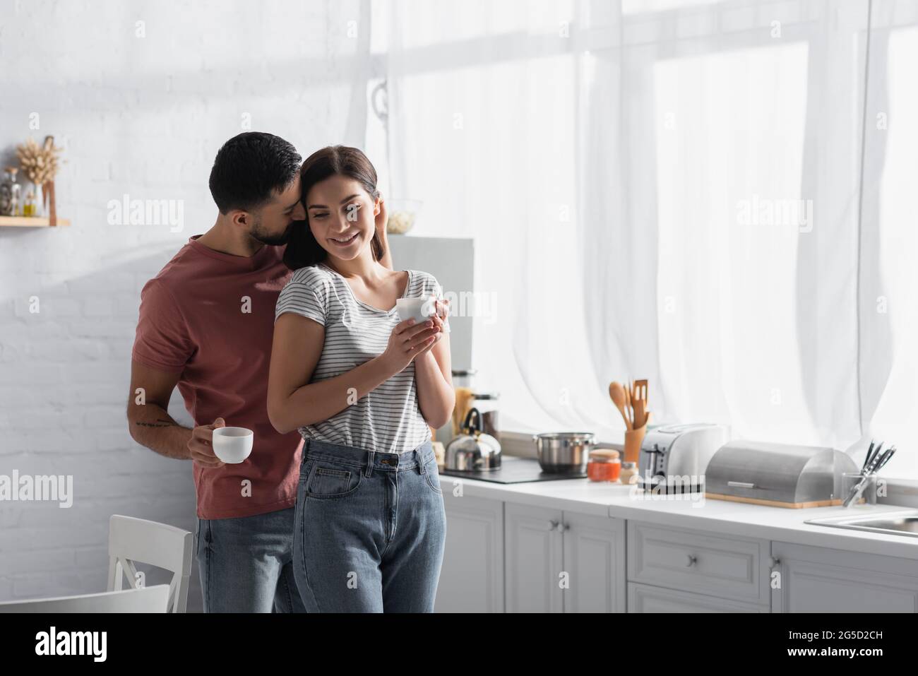 happy young couple hugging and kissing with coffee cups in kitchen Stock Photo
