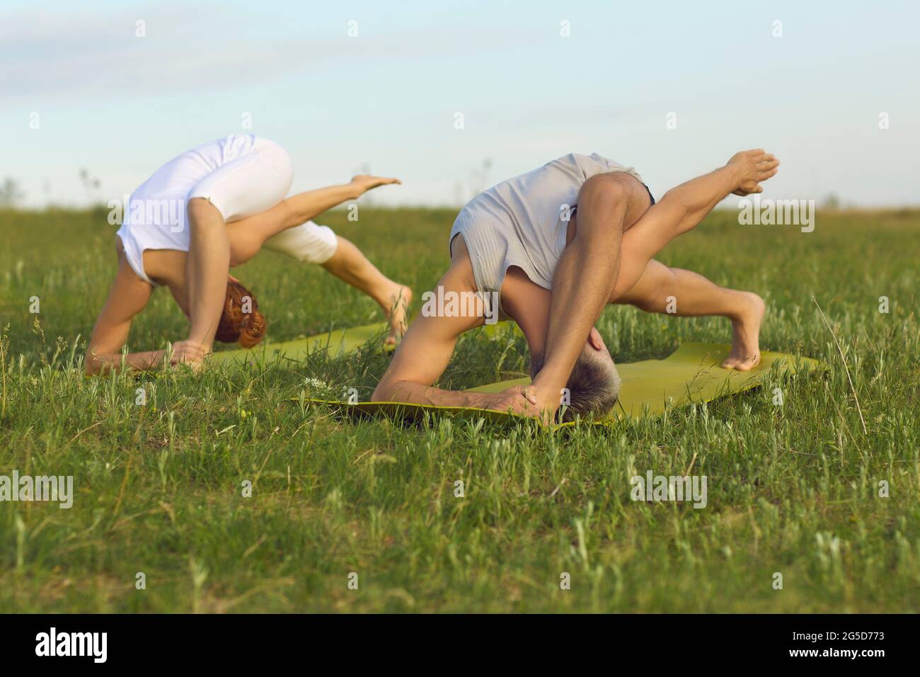 Man and woman doing complicated bending yoga exercises on green grass of summer field Stock Photo