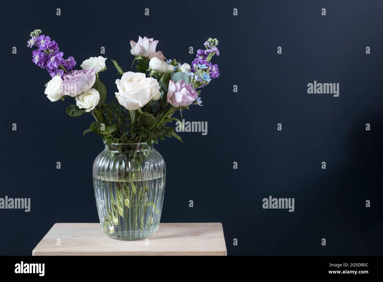 Bouquet of hackelia velutina, purple and white roses, small tea roses, matthiola incana and blue iris in glass vase opposite on black background. Stock Photo