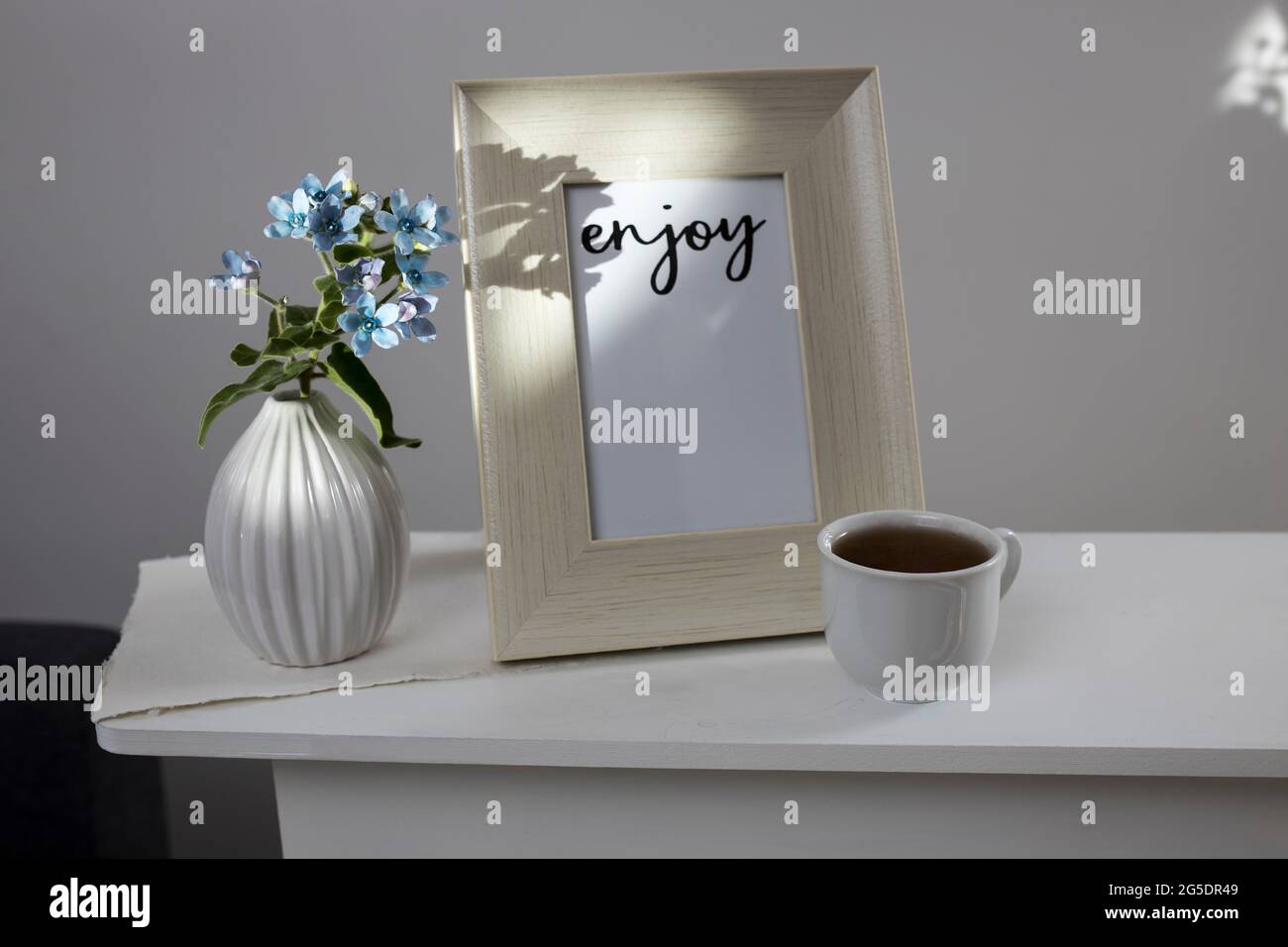 Flower hackelia velutina in a white fluted vase in the style of the seventies, a photo frame with the words Enjoy, and a cup of coffee on the dresser. Stock Photo