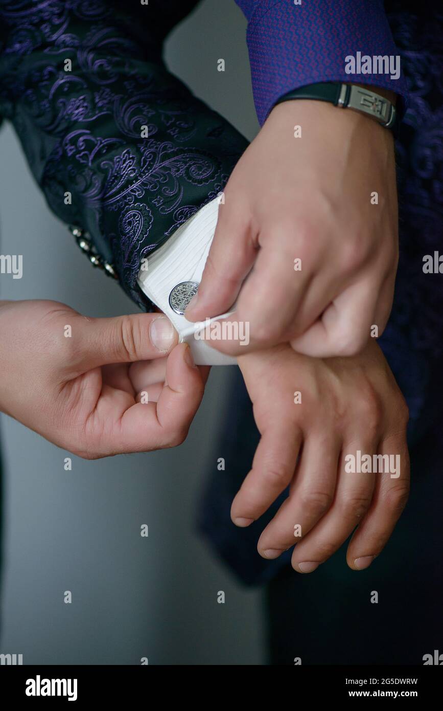 Caucasian young man adjusting silver cufflink of groom wearing dark blue silky tuxedo. Best man helping the groom with the elegant attire. Stock Photo