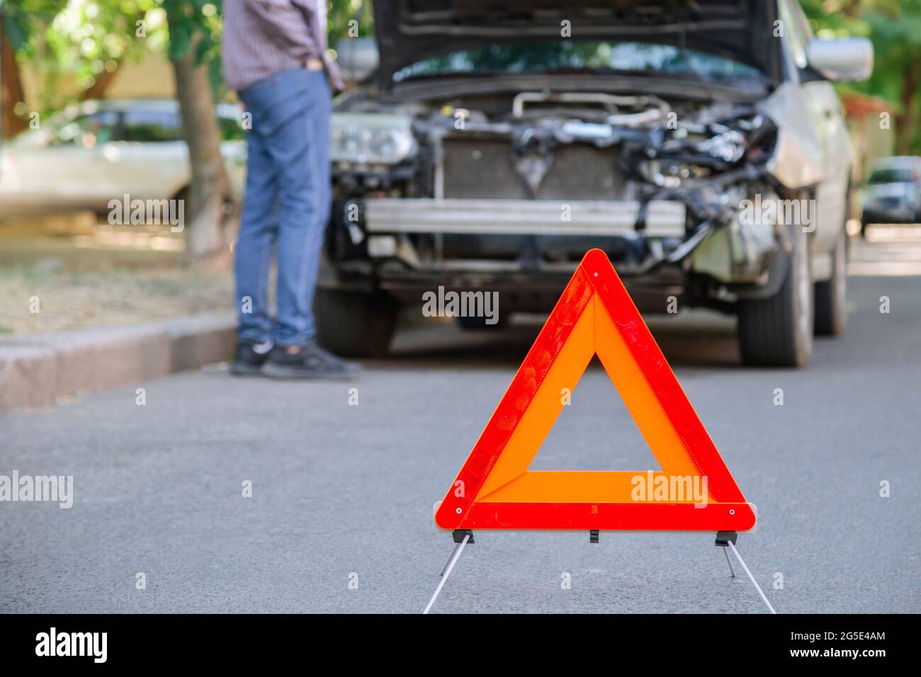 Red triangle warning sign of car accident on road in front of wrecked car. Man inspecting wrecked car after accident. Driver look under the hood of Stock Photo