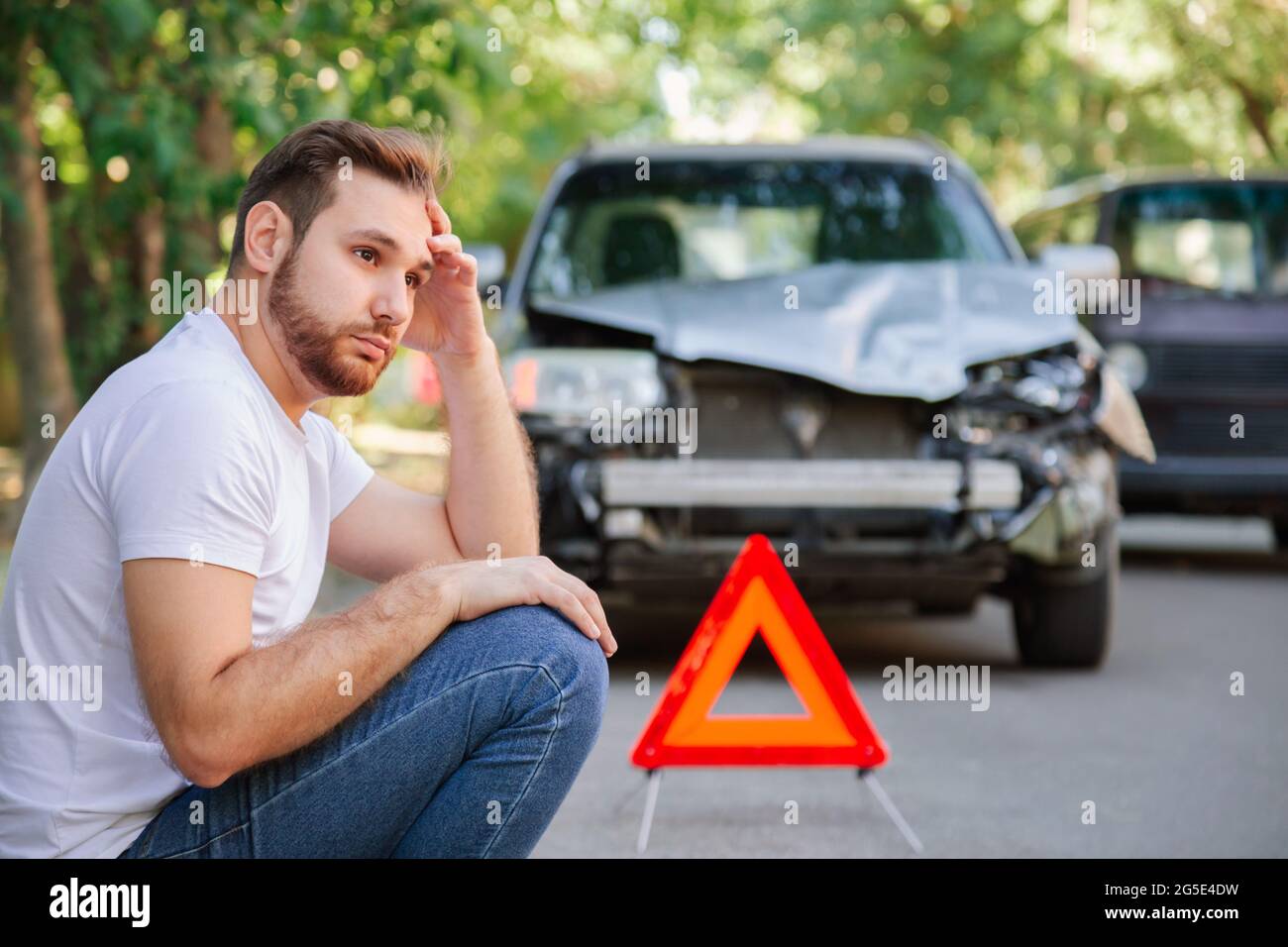 Portrait of upset man sitting on road near wrecked car after car accident with red triangle. Caucasian scary driver man sitting near his wrecked car Stock Photo