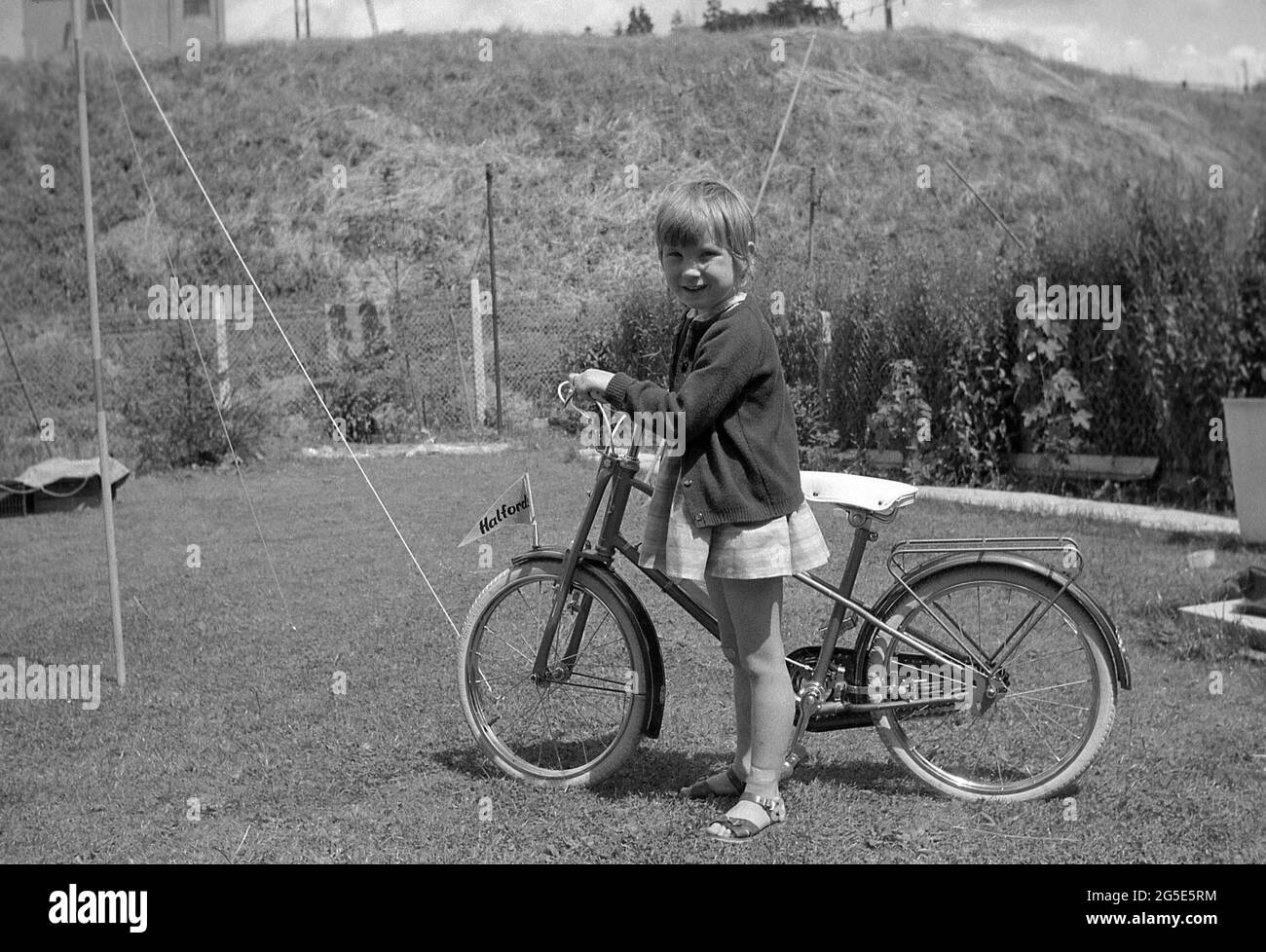 1960s, historical, a young girl in a garden standing proudly with her bicycle, England, UK. On the front mudguard of the bicycle is a small flag with the name Halfords, a leading bicycle retailer of the era. Stock Photo