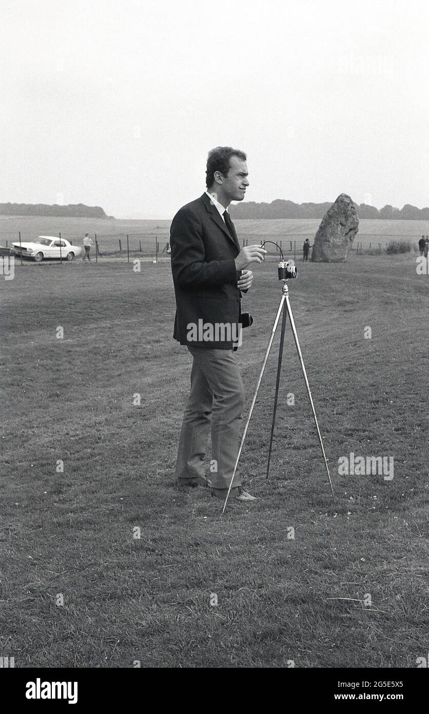 1960s, historical, a man in a jacket and tie using a camera on a tripod to take a picture of the ancient stones at Stonehenge, Wiltshire, England, UK. At this time one could walk freely around the prehistoric stones but in the following decade, in 1977, due to the increasing large numbers of visitors, damage to the grass and general erosion at the sacred, ancient burial site, the stones were roped off preventing access. On land owned at one time by King Henry VIII, the Amesbury Abbey Estate, Cecil Chubb brought the plot and the stones at an auction in 1915 and in 1918 gave it to the nation. Stock Photo