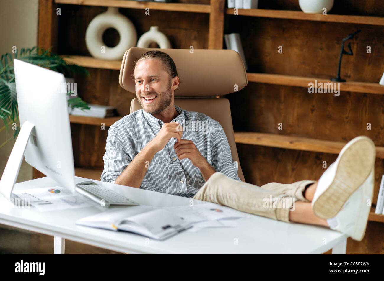 Attractive positive young adult american man, manager or ceo, sitting in office with feet on table, using computer, communicating via conference call with colleagues or friends, reading news, smiling Stock Photo