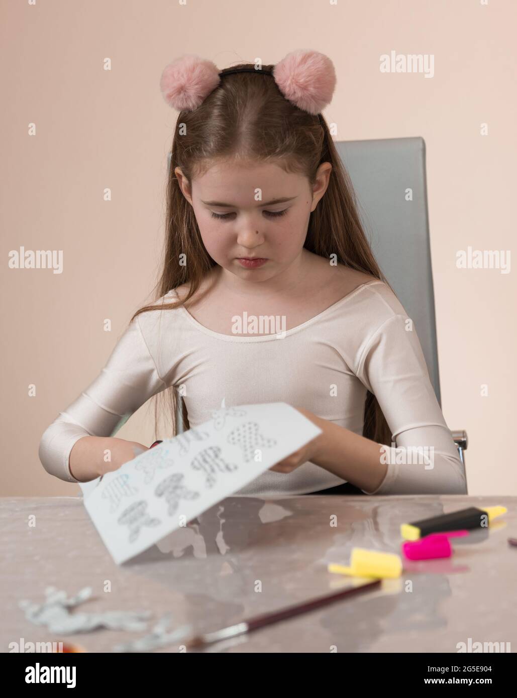 A school-age girl is sitting at a table and is concentrating on cutting jewelry and gifts out of paper with stationery scissors. Long hair and a headb Stock Photo