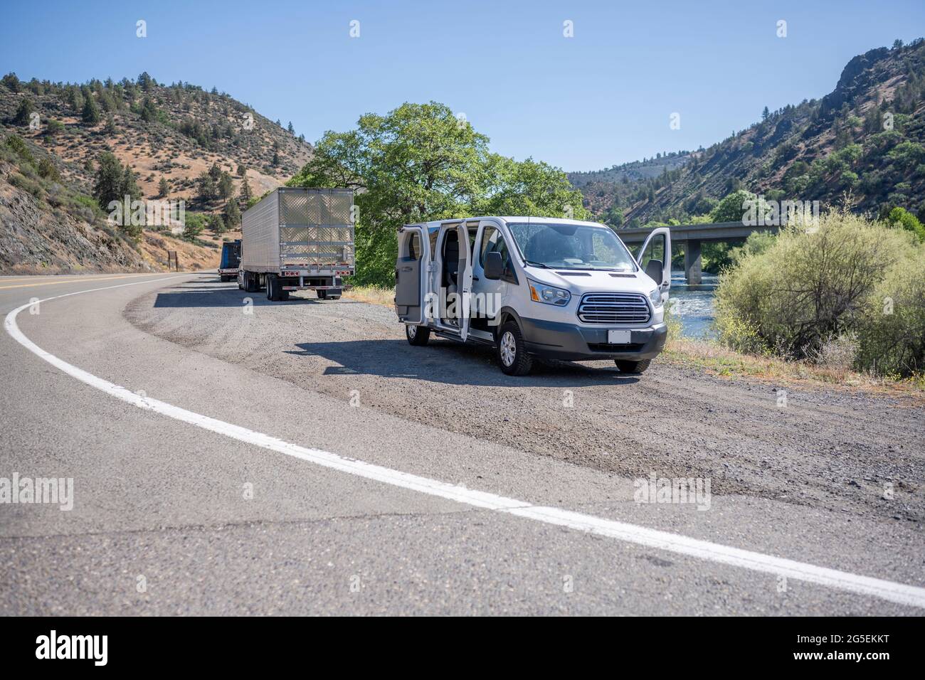 Commercial compact mini van and loaded big rig semi trucks with semi trailers take a break standing on the road shoulder on the bank of a mountain riv Stock Photo