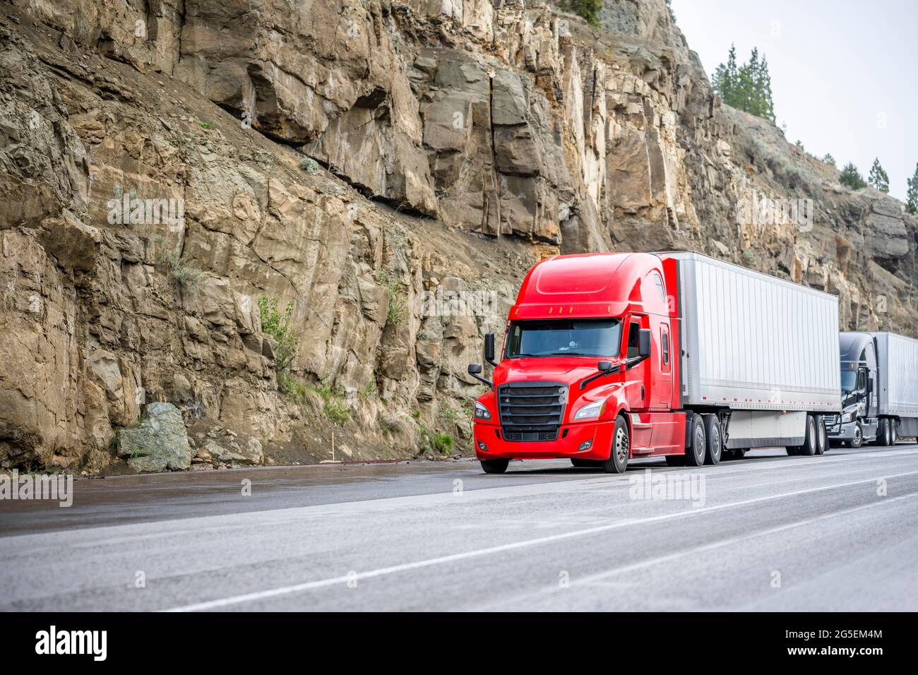 Group of big rigs semi trucks tractors transporting cargo in different semi trailers standing off road in a line near a stone cliff take a break at th Stock Photo