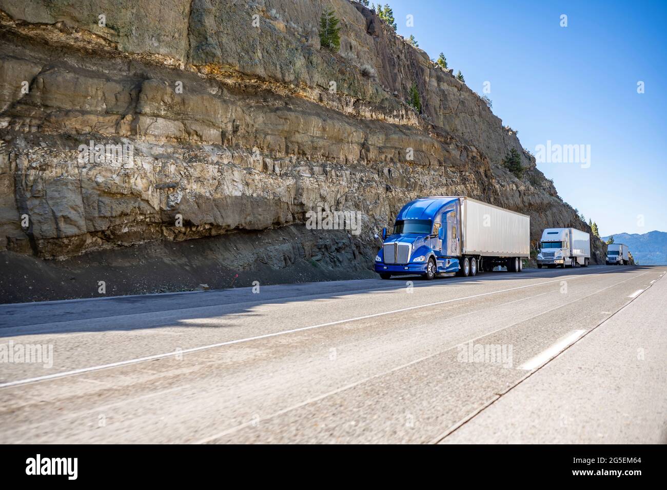 Group of big rigs semi trucks tractors transporting cargo in different semi trailers standing off road in a line near a stone cliff take a break at th Stock Photo