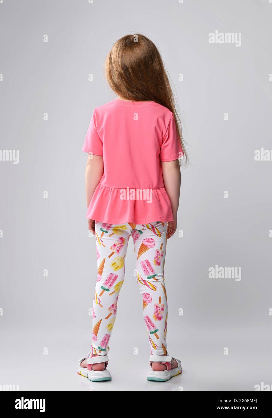 Red-haired kid girl in pink t-shirt, colorful pants and sandals stands still with her back to camera Stock Photo