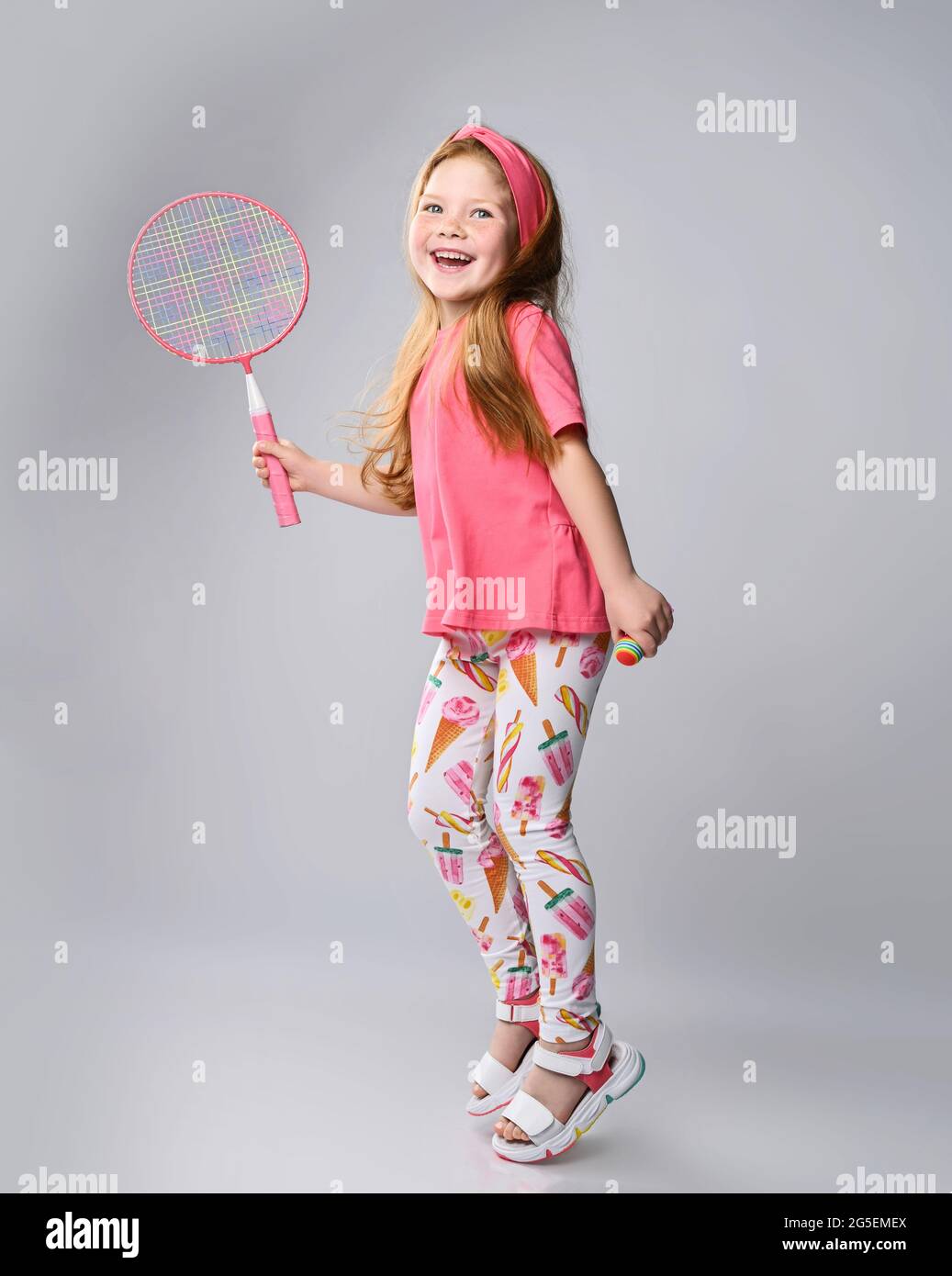 Active, laughing red-haired kid girl in pink t-shirt and headband jumps holding badminton racquet in hand, plays outdoor Stock Photo