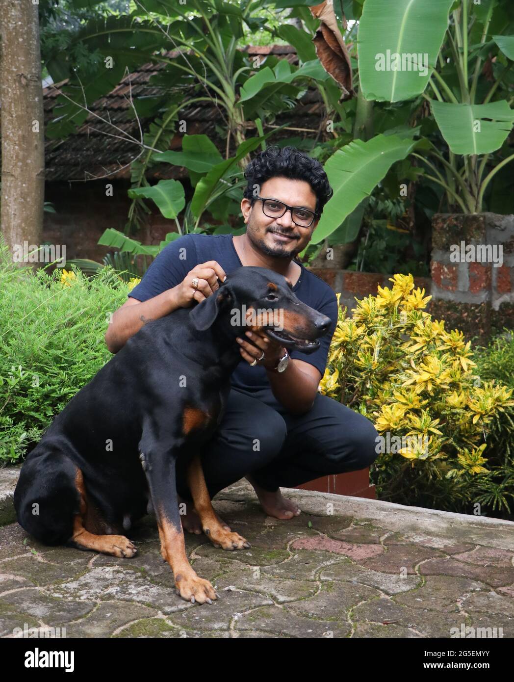 a handsome brown guy casually squatting on the ground petting his black-brown doberman pinscher dog in a garden Stock Photo
