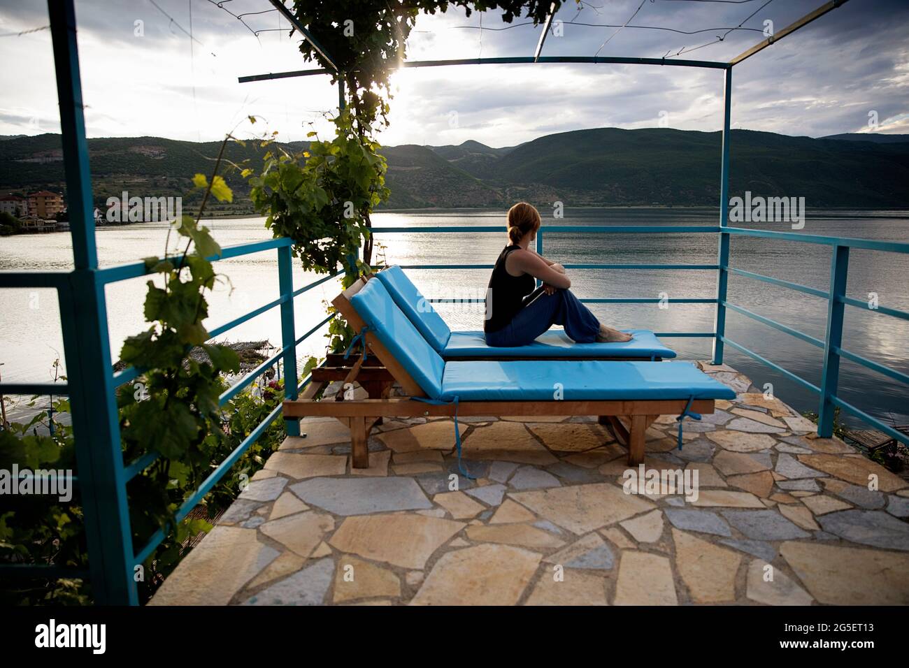Woman in casual clothing sitting on wooden deck chair on a terrace looking to the lake at sunset, Lin, Lake Ohrid, Korca region, Albania Stock Photo