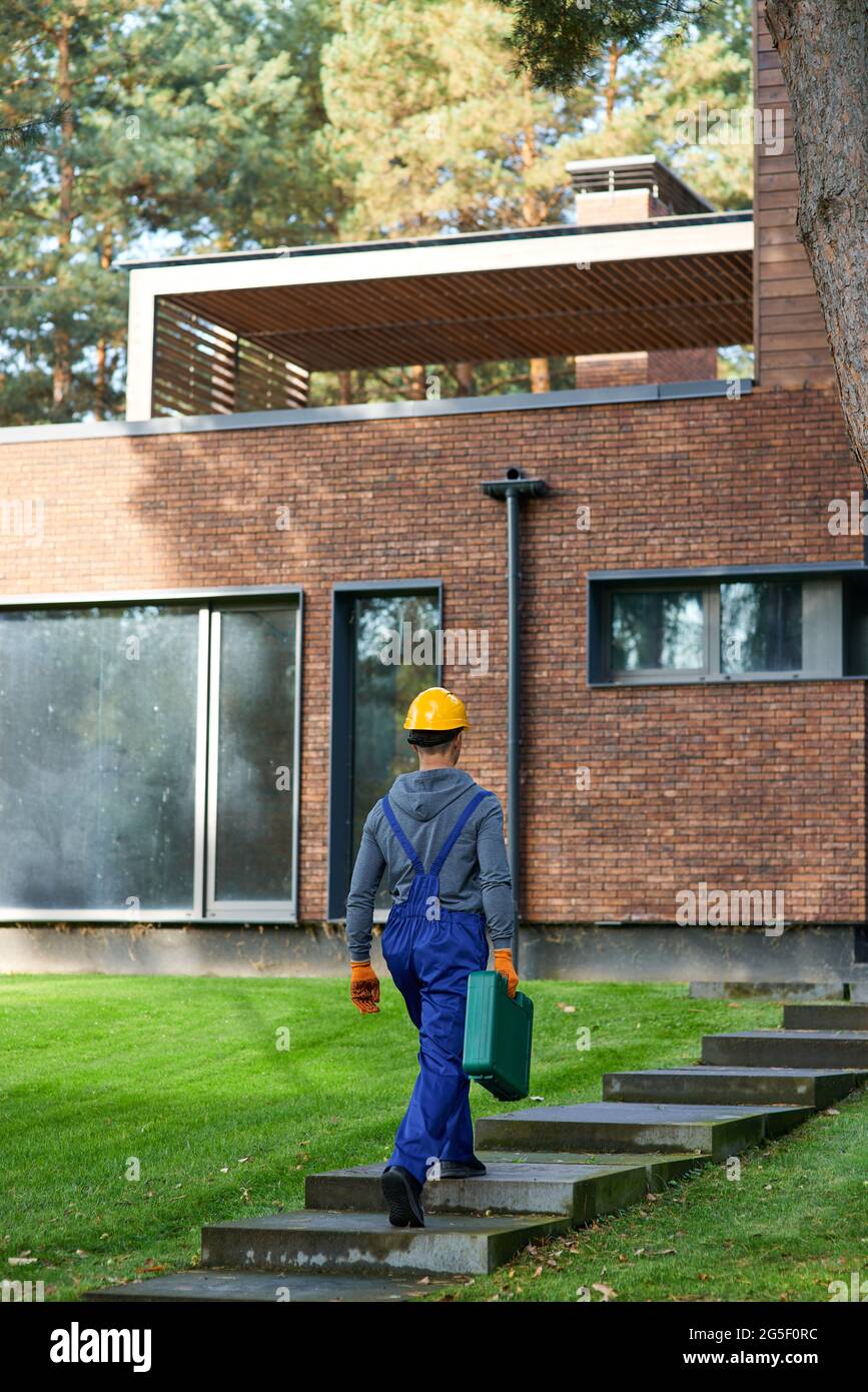 Full length shot of young male builder in blue overalls carrying toolbox at cottage construction site Stock Photo