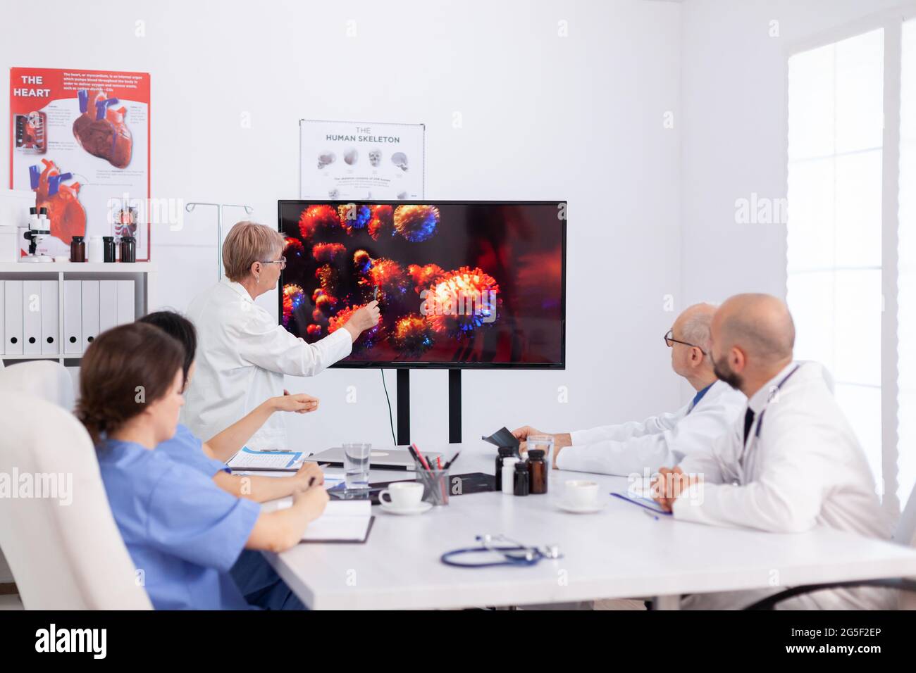 Senior practitioner doctor woman pointing virus sickness on monitor in hospital meeting room discussing with coworkers during brainstorming. Medical team of physicians planning vaccine against coronavirus Stock Photo