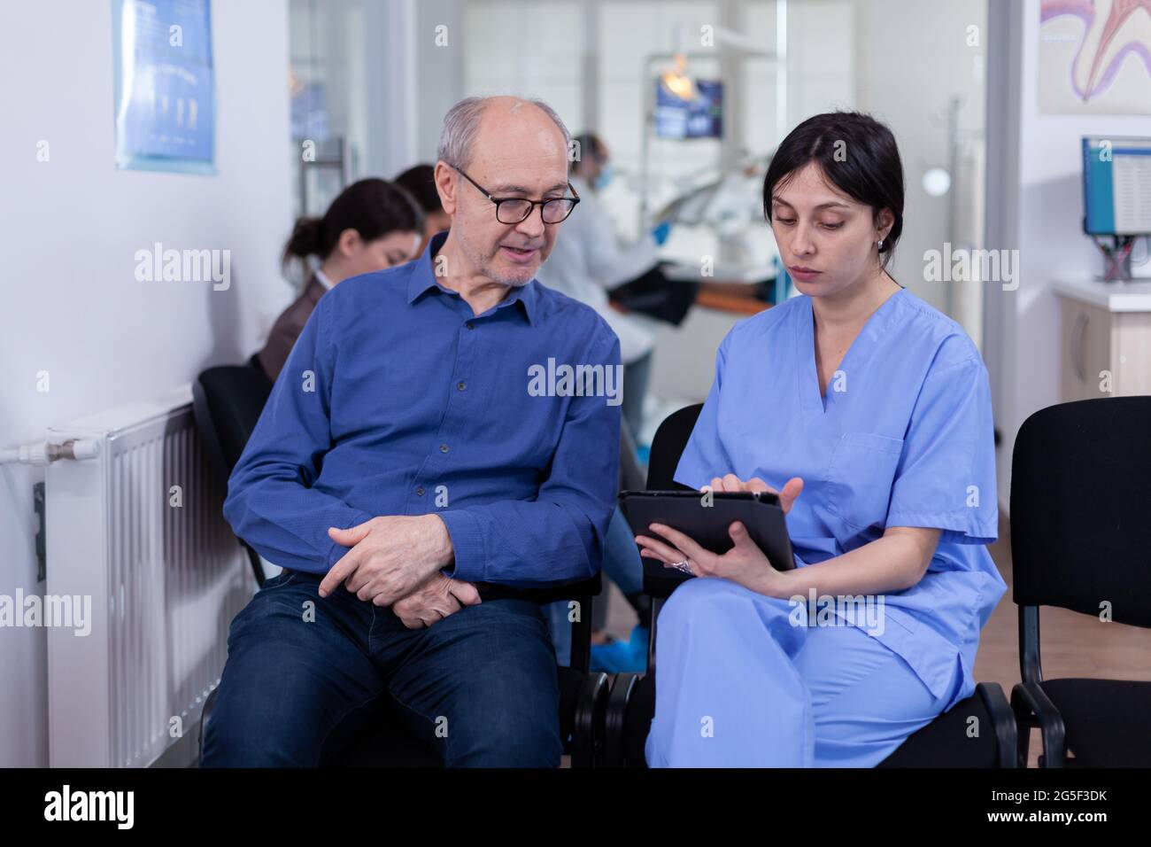 Nurse taking notes on tablet pc discussing with senior patient in dental office, explaining diagnosis. Assistant filling writing healthcare form. Dentistiry hospital. Stock Photo