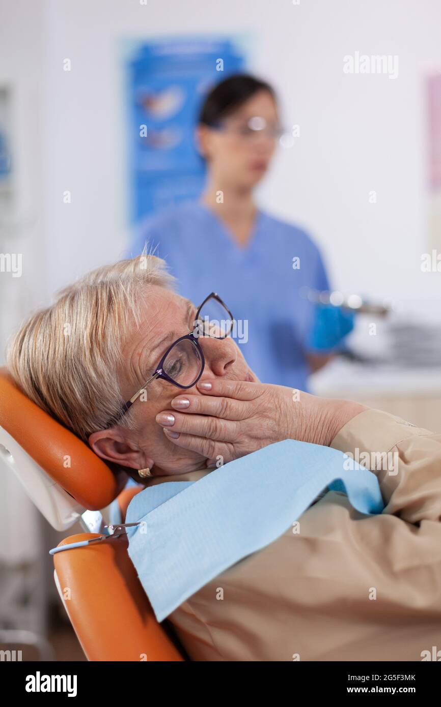 Middle age patient touching mouth with painful expression sitting on chair in dentist cabinet. Senior woman in healthcare hospital accusing and complaining about tooth. Stock Photo