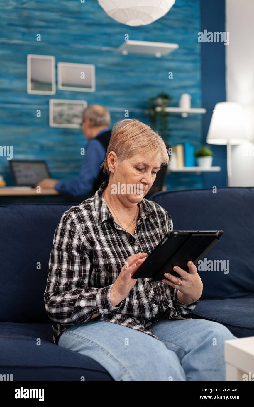 Middle aged lady relaxing on sofa reading on tablet pc anjoying content. Elderly woman using moder technoloy tablet pc in home living room and husband working on laptop computer Stock Photo