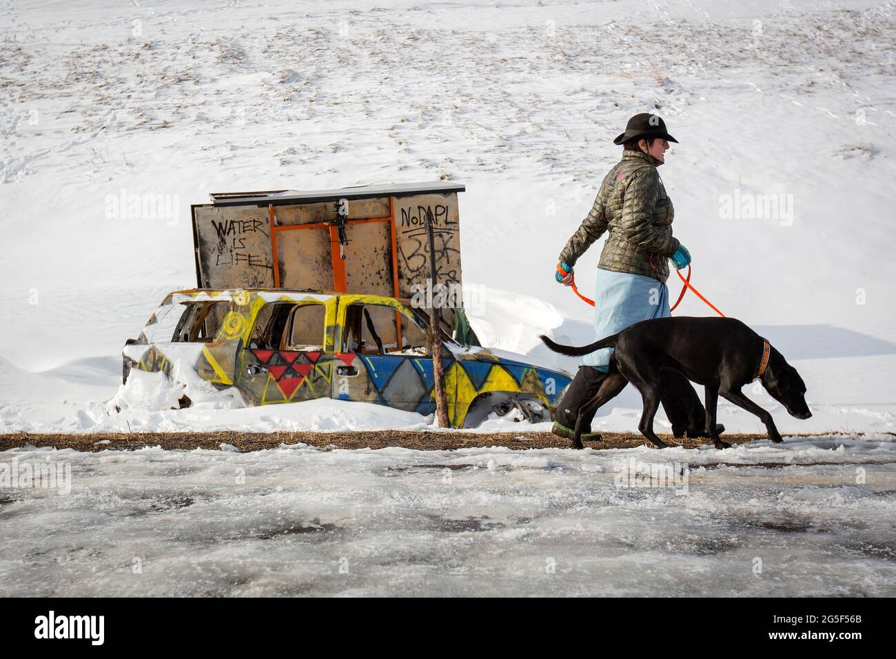 En demonstrant går tur med hunden sin forbi en av de utbrente DAPL- bilene som står igjen ved veisperringen der sammenstøtene mellom politi og demonstrater tok sted. Etter måneder med protester klarte Sioux-indianerne ved Standing Rock og deres støttespillere å stoppe US Army Corps of Engineers fra å bygge den omstridte DAPL - Dakota Access Pipeline - under relven som går rett gjennom Dakota-indianernes reservat. Aksjonene fikk også gamle motsetninger mellom indianerne og norskamerikanerne til overflaten. Stock Photo