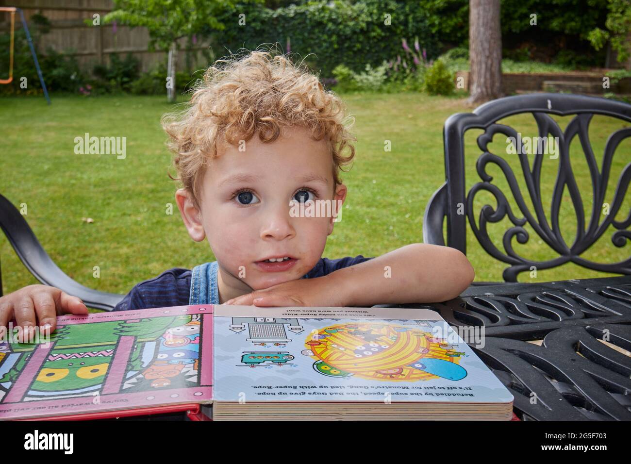 A white Caucasian small boy with curly blond hair age 2 -3 sits facing the camera with a child's book at a table in a garden on a sunny day in England A white Caucasian small boy with curly blond hair age 2 -3 sits facing the camera with a child's book at a table in a garden on a sunny day in England