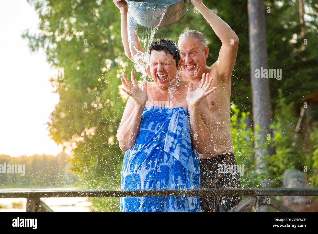 Aged couple having fun after Finnish sauna on wooden cottage pier in a lake. Mature man pouring cold water over his wife. Typical Finnish summer. Stock Photo