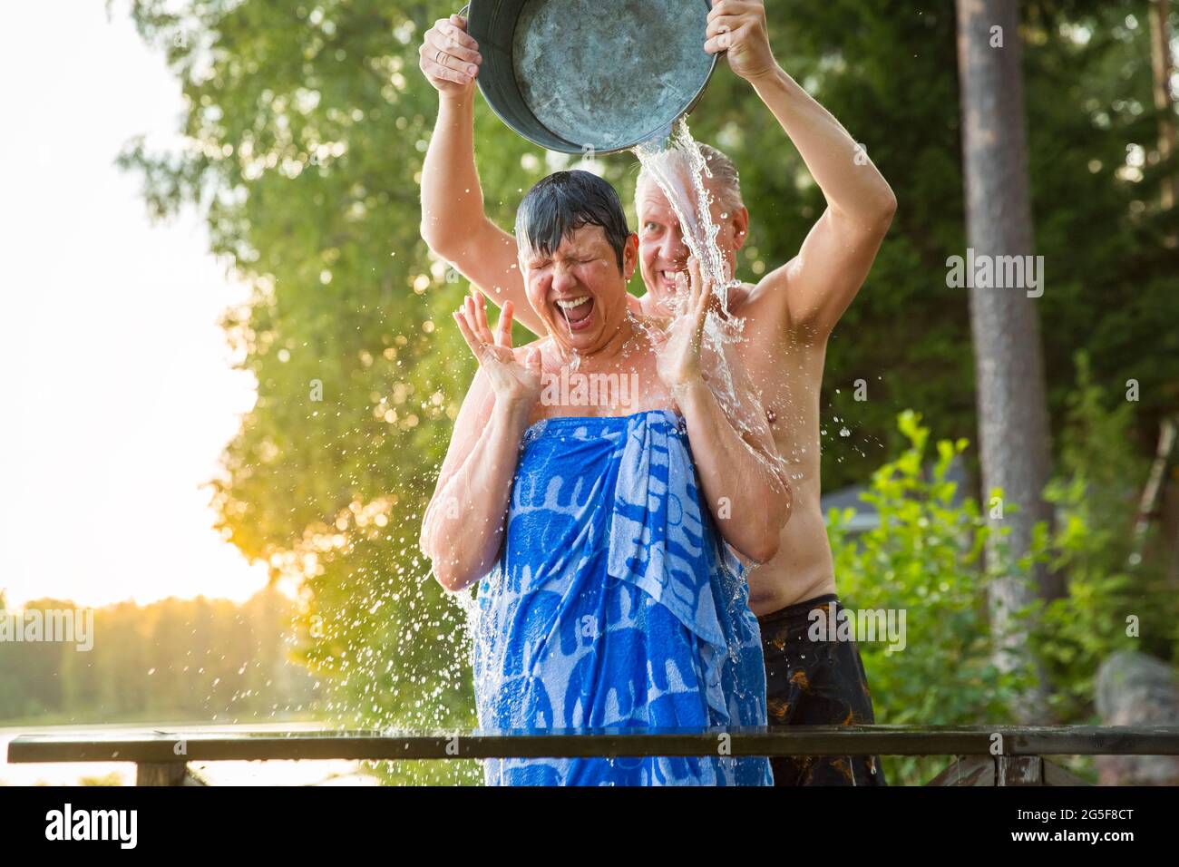 Aged couple having fun after Finnish sauna on wooden cottage pier in a lake. Mature man pouring cold water over his wife. Typical Finnish summer. Stock Photo