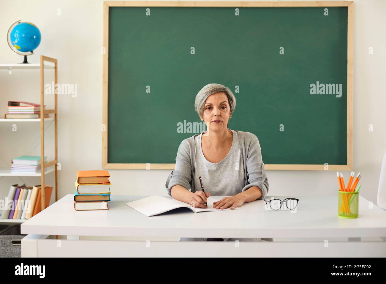 Teacher teaches. Serious senior woman teacher looking at the camera at lesson on the background of a greenboard in the classroom. Stock Photo