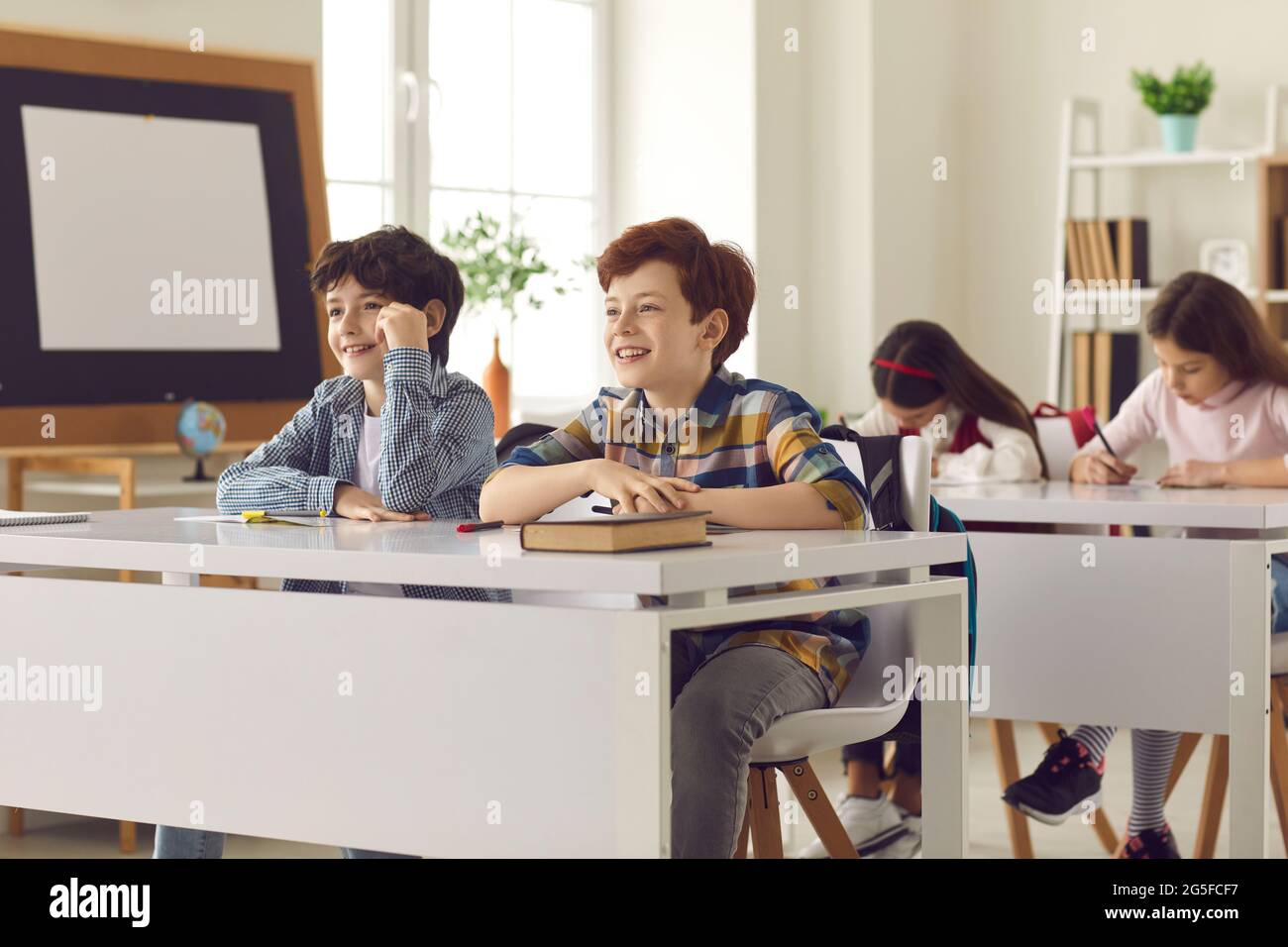 Happy elementary school boys listening to teacher sitting at desk in modern classroom Stock Photo