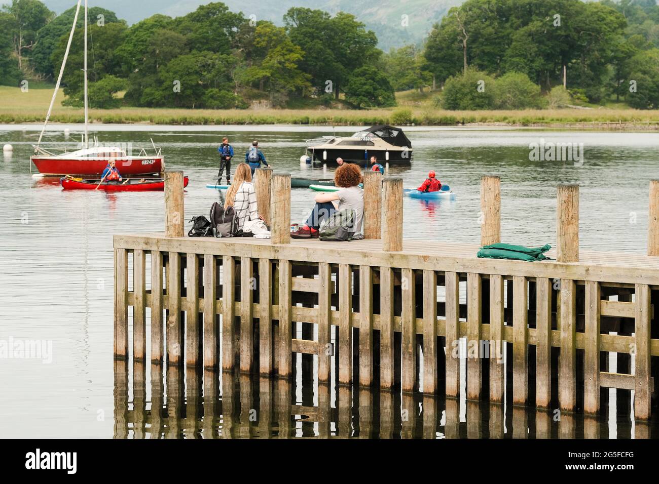 Two people sat on a wooden jetty at Ambleside watch activity on Lake Windermere in the Lake District, Cumbria, UK. Stock Photo