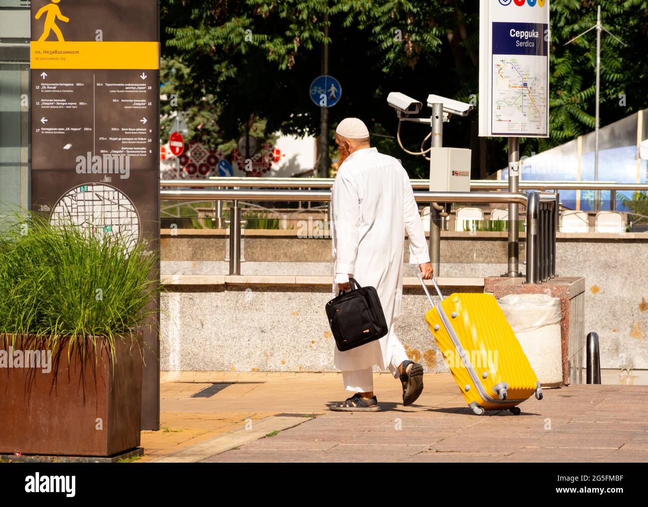 Middle age muslim man wearing white thawb robe and kufi cap with a lapton case pulling yellow suitcase in the streets of Sofia, Bulgaria Stock Photo