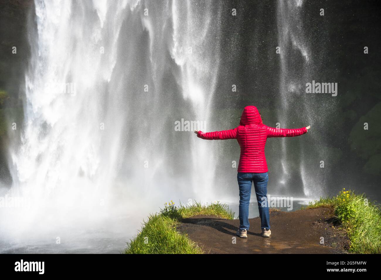 Woman with outstrtched arms at the foot of a majestic waterfall on a sunny summer day. Power in nature. Stock Photo