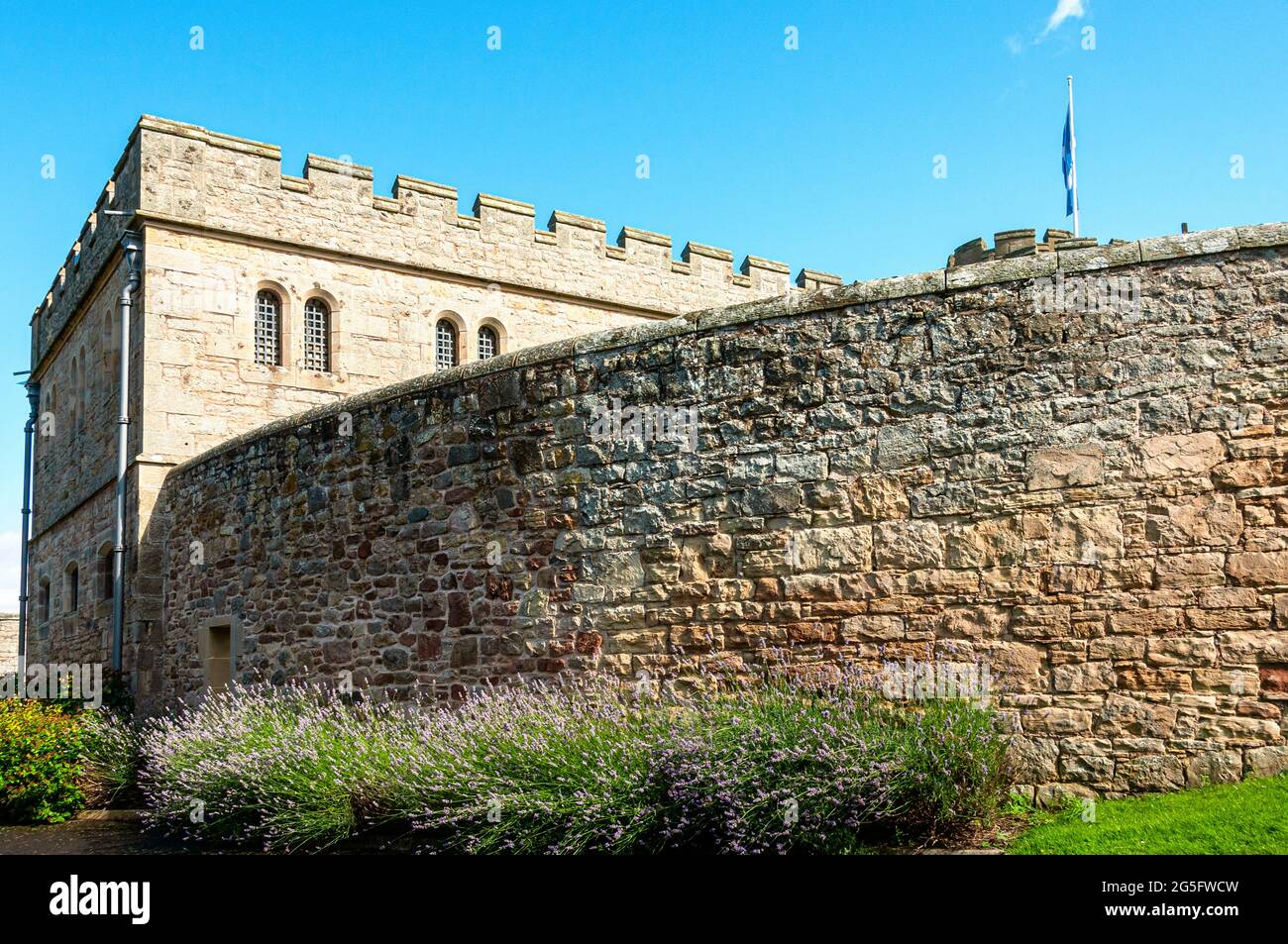 A two storey cell block of Jedburgh Prison flying the Saltire from a round tower in bright sunshine is partly hidden by a high wall of dressed stone Stock Photo