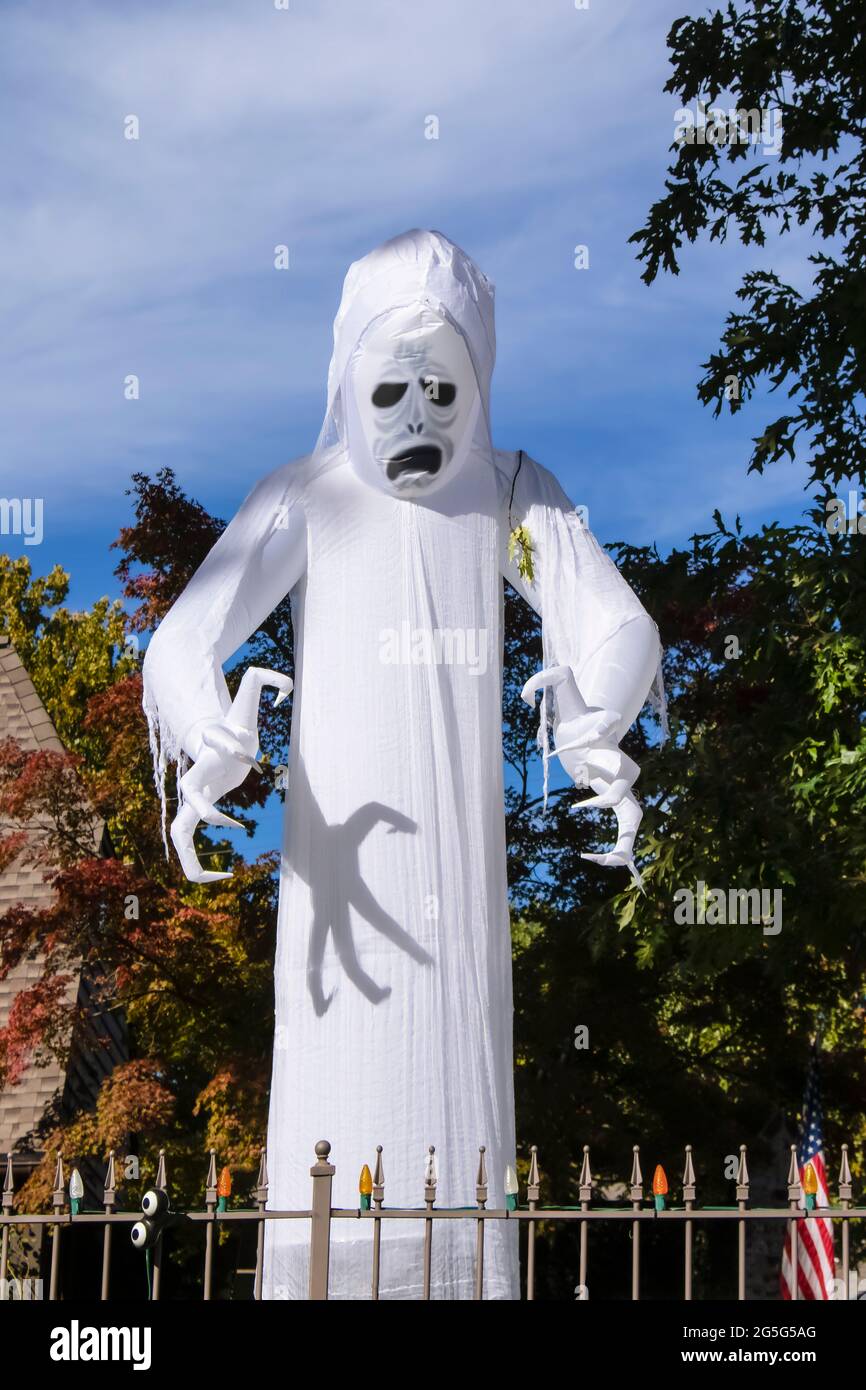 Scary giant white ghost - zombie blowup Halloween decoration towers above house surrounded by Autumn trees with claws ready to attack behind fence wit Stock Photo