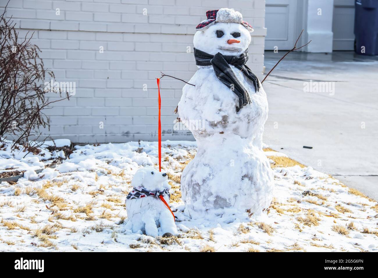 Snowman with coal for eyes and plaid cap with earflaps and scarf holding a snow dog with kerchief and ball by leash Stock Photo