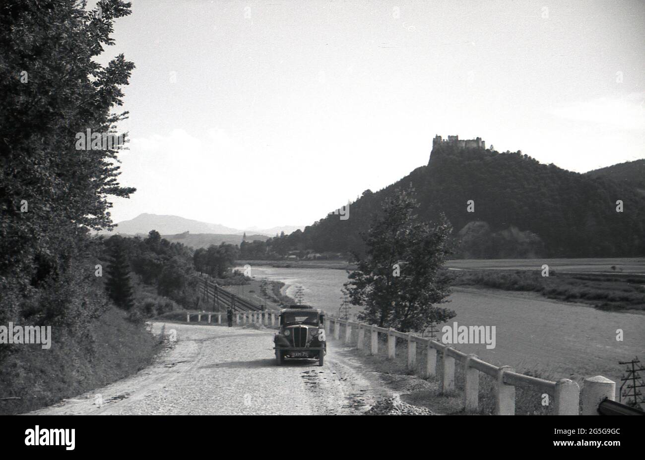 1930s, hisotrical, touring Czechoslavia pre-ww2, a British car, a Morris 8, parked on a rural gravel road, beside a rail track and river, with a castle on a hill in forest in the background. This area of Czechoslovakia, was known and referred to at this time as the Sudetenland, as they were many ethnic German people living in the western Czech lands of Bohemia. Stock Photo