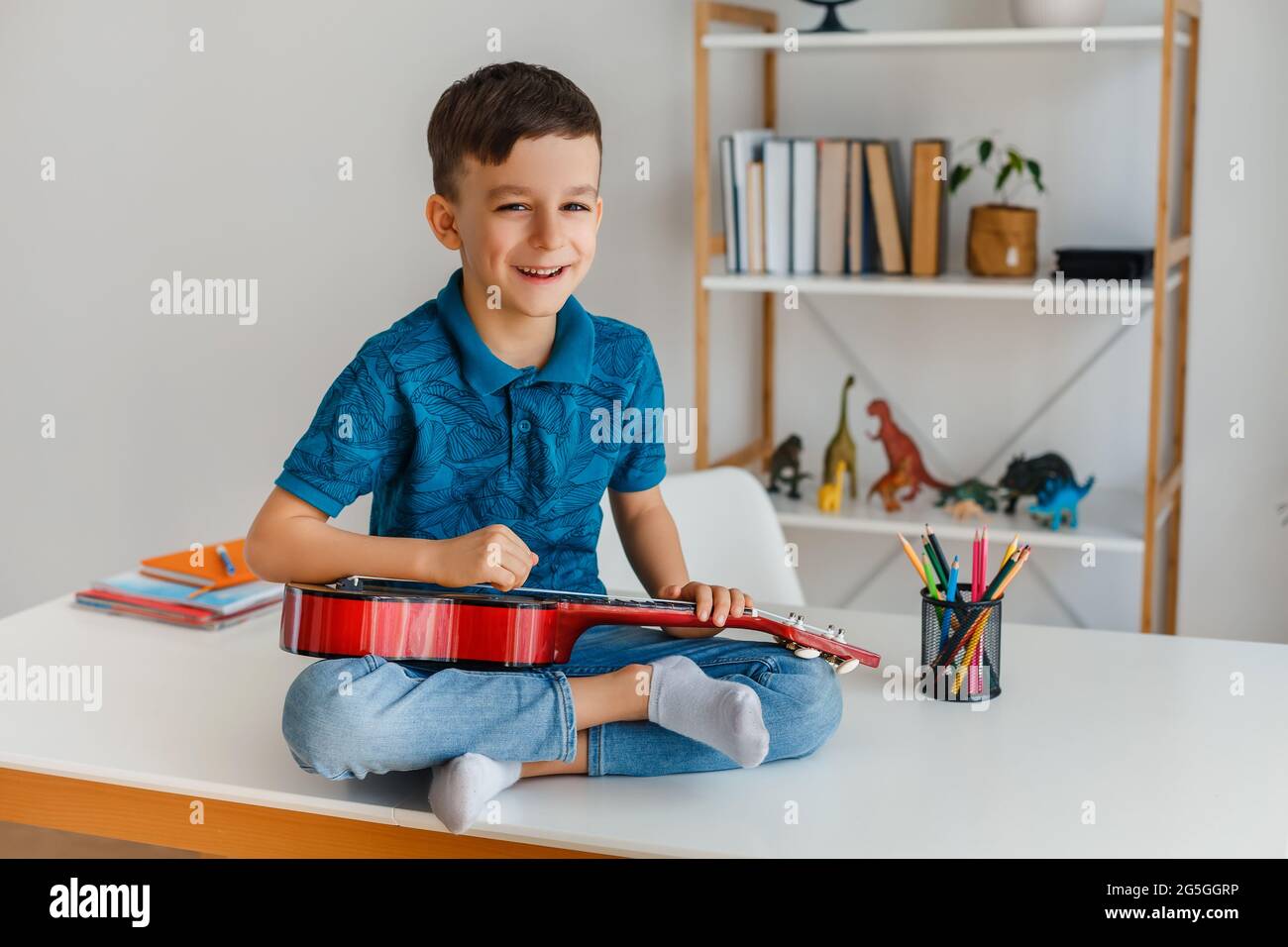 Talented kid playing soprano ukulele sitting on desk. Preschool boy learning guitar at leisure. Concept of early childhood education and music hobby Stock Photo