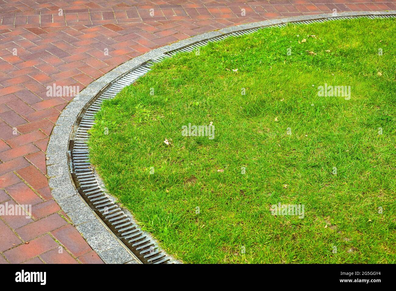 curved iron grid drainage system in the park by the green lawn meadow and walkway of red paving slabs with copy space, nobody. Stock Photo