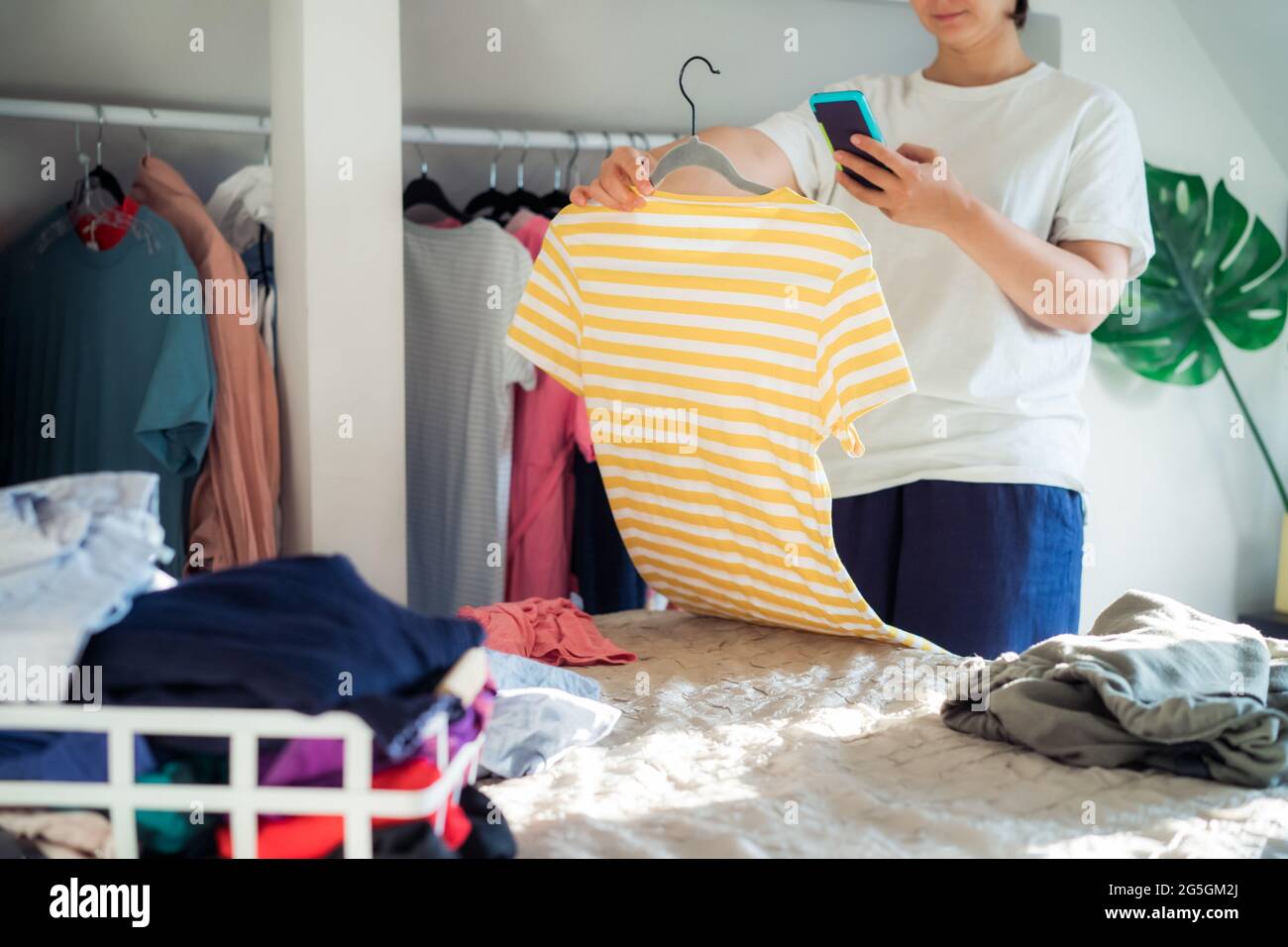 Woman taking photo of t-shirt using her smartphone for selling or donating her clothes. Decluttering Clothes, Sorting, And Cleaning Up. Reuse, second Stock Photo