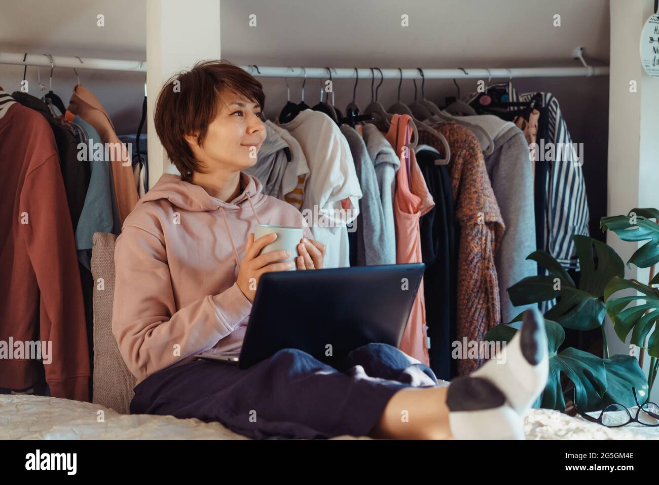 Woman with cup of coffee sitting on the bed and using laptop and enjoy the moment. Apartment with an open wardrobe. Cozy home. Online communication Stock Photo