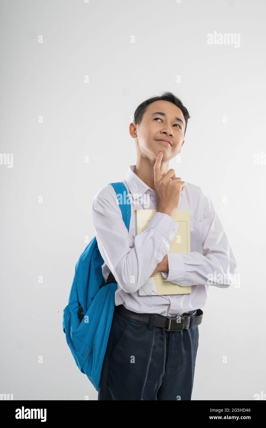 a boy in junior high school uniform smiling looking up when thinking gesture when holding a book and a backpack with copyspace Stock Photo