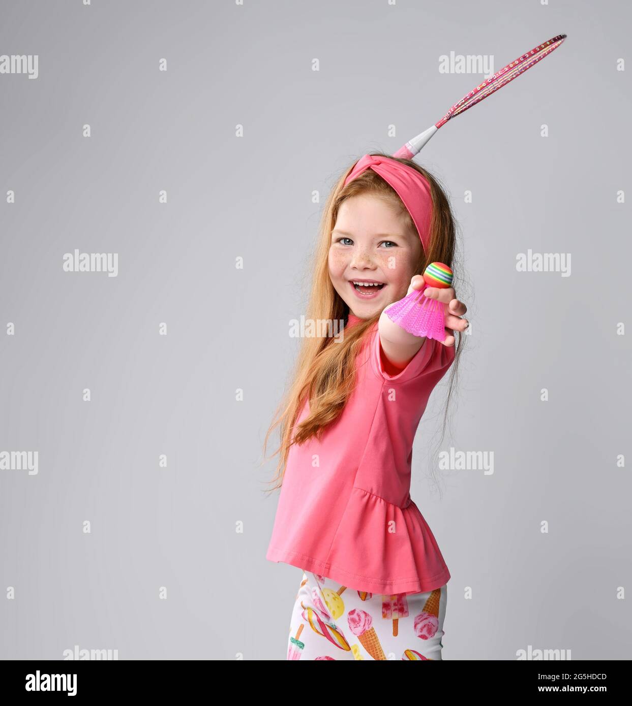 Laughing red-haired kid girl in pink t-shirt and headband plays badminton, holds shuttlecock in hand, swings racquet Stock Photo