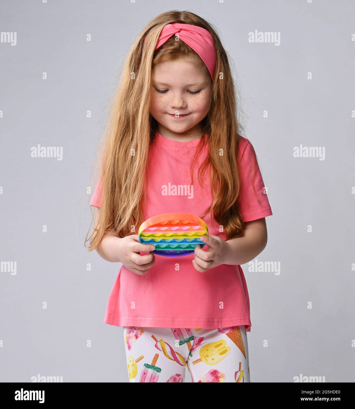 Happy kid girl in pink t-shirt and headband plays a new trendy sensory rainbow color round toy - pop it, pushes Stock Photo