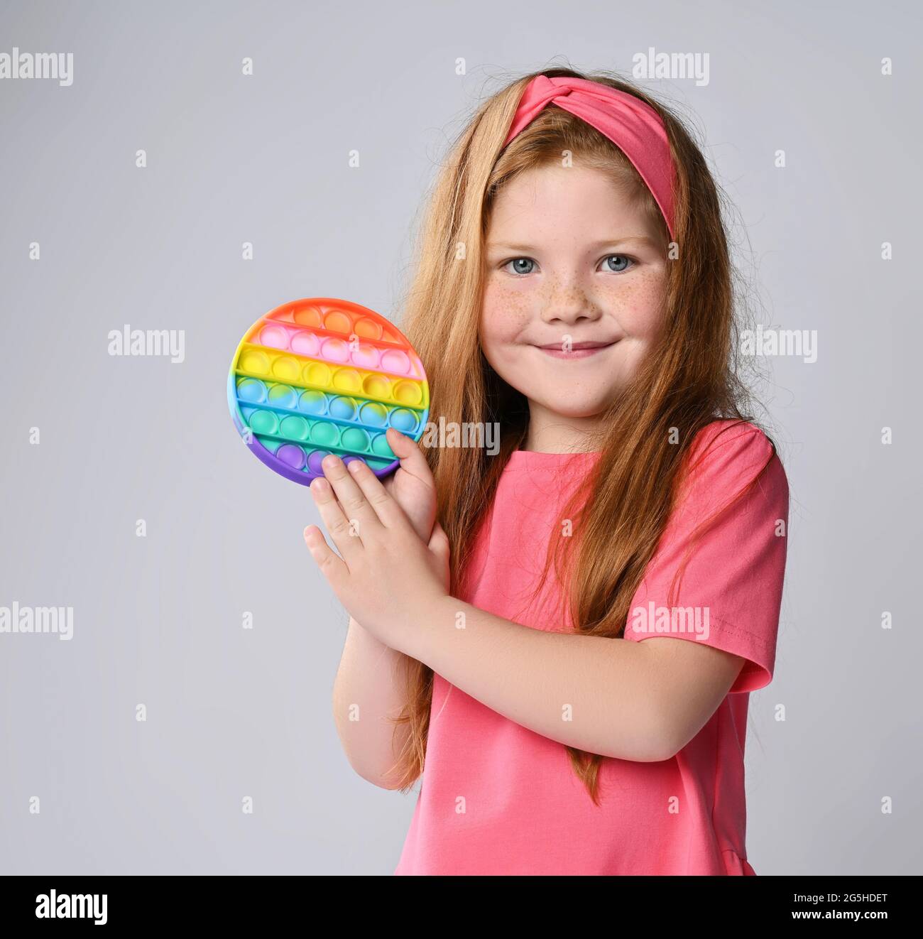 Happy kid girl in pink t-shirt and headband demonstrates, shows her new trendy sensory toy, rainbow color round - pop it Stock Photo