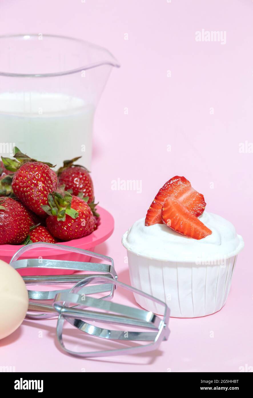 Cupcake with cream and strawberry slices, beaters from the mixer, egg, milk on a pink background. Selective focus. Stock Photo