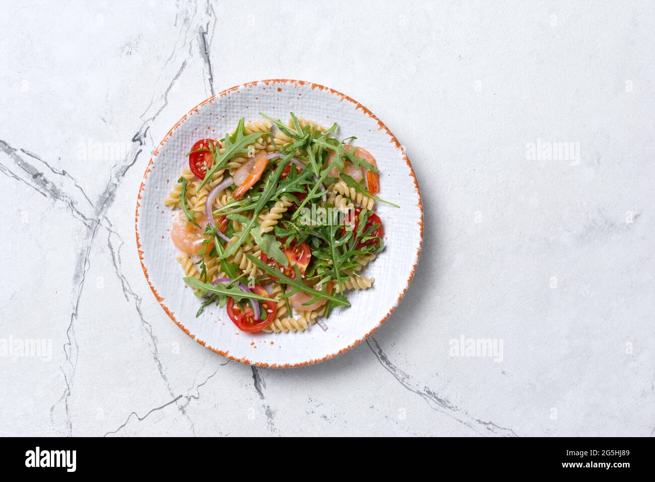 Top view of tasty pasta with shrimps, cherry tomatoes, onion, sesame and green arugula leaves on white marble background Stock Photo