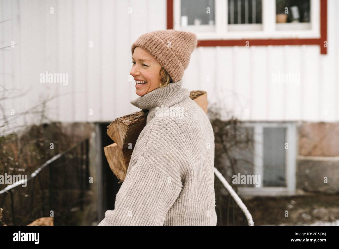 Smiling woman wearing knit hat carrying firewood while looking away Stock Photo
